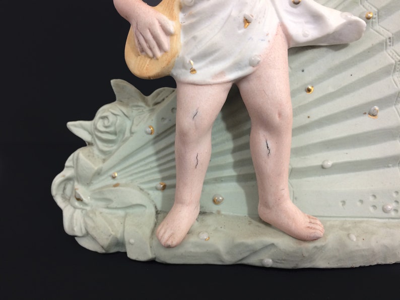 Puede incluir: Una figurita de porcelana de un querub&iacute;n tocando un instrumento musical, de pie sobre una base en forma de abanico verde y blanco con detalles de perlas.