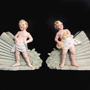 Puede incluir: Dos figuritas de porcelana de querubines, cada una sosteniendo un instrumento musical, se colocan sobre una base verde en forma de abanico. Los querubines est&aacute;n desnudos con una tela blanca drapeada alrededor de sus cinturas. Las bases est&aacute;n decoradas con acentos dorados y puntos blancos.