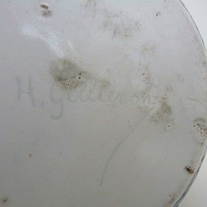 May include: Close-up of the bottom of a white ceramic object with a blue and white design. The bottom is marked with the handwritten signature "H. Gebler"
