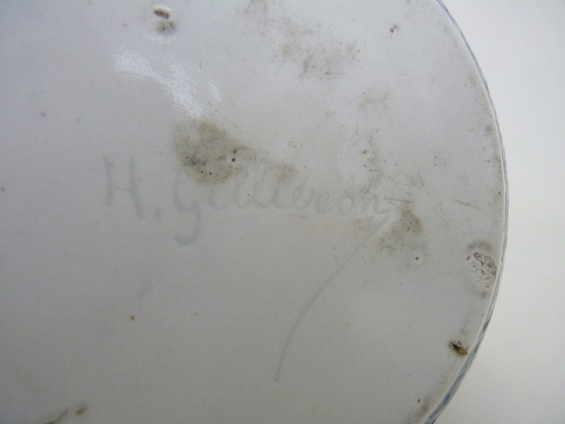 May include: Close-up of the bottom of a white ceramic object with a blue and white design. The bottom is marked with the handwritten signature "H. Gebler"