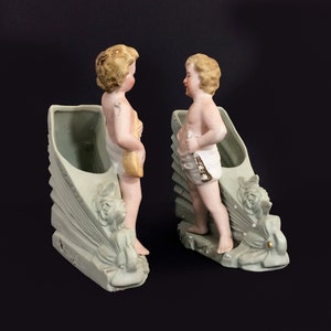 Puede incluir: Dos figuras de querubines de porcelana se encuentran en un par de macetas de cer&aacute;mica verde y blanca. Cada maceta tiene forma de concha con un dise&ntilde;o floral decorativo. Los querubines est&aacute;n desnudos y tienen el pelo rubio.