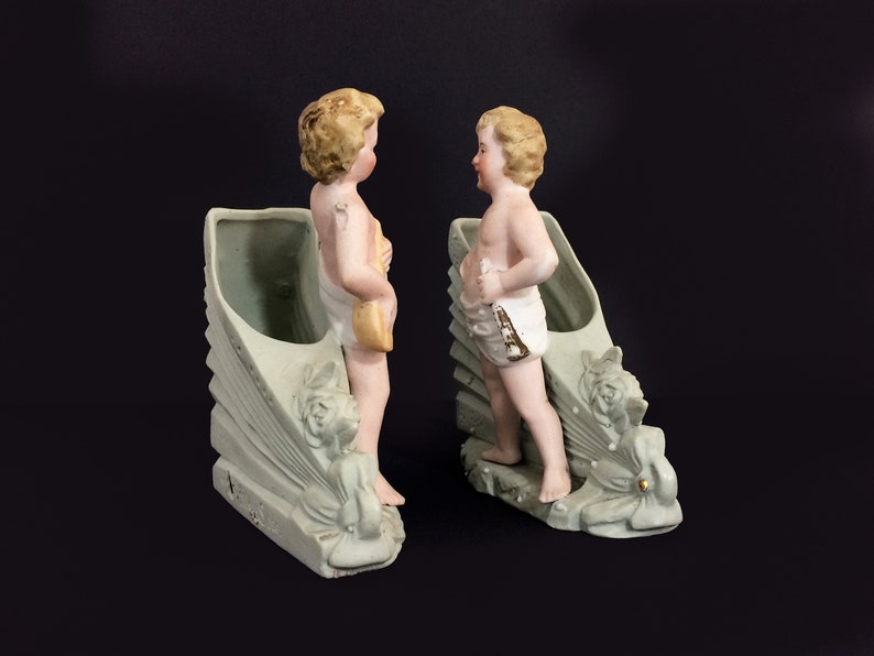 Puede incluir: Dos figuras de querubines de porcelana se encuentran en un par de macetas de cer&aacute;mica verde y blanca. Cada maceta tiene forma de concha con un dise&ntilde;o floral decorativo. Los querubines est&aacute;n desnudos y tienen el pelo rubio.