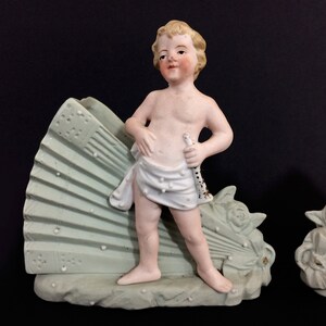 Puede incluir: Par de figuritas de porcelana vintage de querubines, cada uno sosteniendo un instrumento musical. Los querubines est&aacute;n de pie sobre una base en forma de abanico con detalles florales. Las figuritas est&aacute;n pintadas en tonos de blanco, rosa y verde.