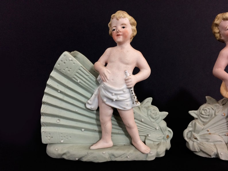 Puede incluir: Par de figuritas de porcelana vintage de querubines, cada uno sosteniendo un instrumento musical. Los querubines est&aacute;n de pie sobre una base en forma de abanico con detalles florales. Las figuritas est&aacute;n pintadas en tonos de blanco, rosa y verde.