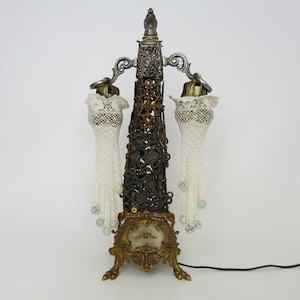 May include: A vintage-style lamp with a gold and silver finish. The lamp has two shades made of white lace with beaded accents. The lamp has a decorative base with intricate details.