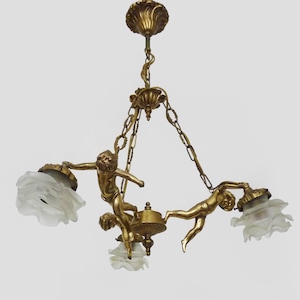 Puede incluir: Lámpara de araña dorada antigua con tres figuras de querubines que sostienen pantallas de vidrio esmerilado. La lámpara tiene detalles ornamentados y un soporte decorativo para el techo, suspendida por cadenas.