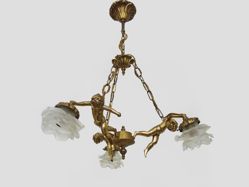 Puede incluir: L&aacute;mpara de ara&ntilde;a dorada antigua con tres figuras de querubines que sostienen pantallas de vidrio esmerilado. La l&aacute;mpara tiene detalles ornamentados y un soporte decorativo para el techo, suspendida por cadenas.