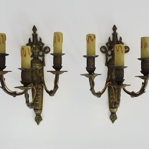 May include: A pair of antique brass wall sconces, each with three candle holders. The sconces feature a decorative design with scrolls and floral motifs.