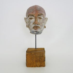 Puede incluir: Una escultura de cerámica de una cabeza humana con una expresión neutral. La escultura está pintada en tonos de marrón, blanco y gris. Está montada sobre una base de madera.