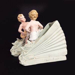 Puede incluir: Un jarr&oacute;n de porcelana blanca con dos figuras de querubines. El jarr&oacute;n tiene forma de abanico y presenta un dise&ntilde;o floral decorativo. Los querubines sostienen instrumentos musicales. El jarr&oacute;n est&aacute; marcado con el n&uacute;mero "12106 S. DEP".