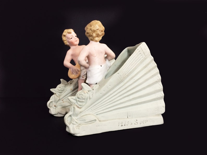 Puede incluir: Un jarr&oacute;n de porcelana blanca con dos figuras de querubines. El jarr&oacute;n tiene forma de abanico y presenta un dise&ntilde;o floral decorativo. Los querubines sostienen instrumentos musicales. El jarr&oacute;n est&aacute; marcado con el n&uacute;mero "12106 S. DEP".
