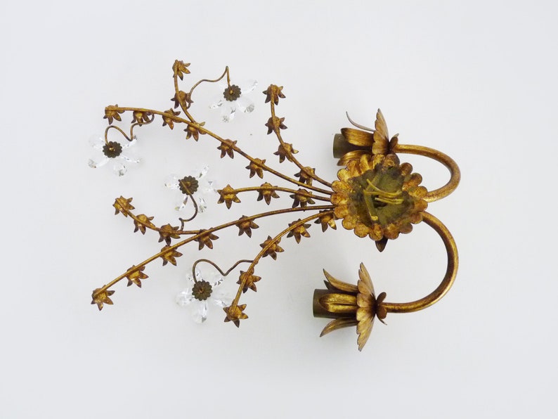 May include: Gold metal wall sconce with a floral design and clear crystal accents. The sconce has two arms that curve upwards and are decorated with leaves and flowers. The sconce is perfect for adding a touch of elegance to any room.