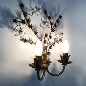 May include: A gold metal wall sconce with a floral design. The sconce has two light bulbs and crystal accents.
