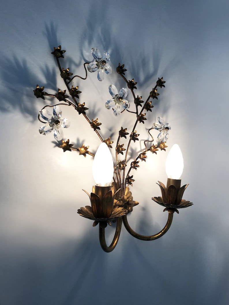 May include: A gold metal wall sconce with a floral design. The sconce has two light bulbs and crystal accents.