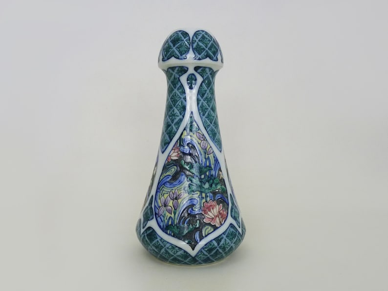 May include: A ceramic bottle stopper with a green, blue, and white floral design. The stopper is shaped like a bottle neck and has a decorative top.