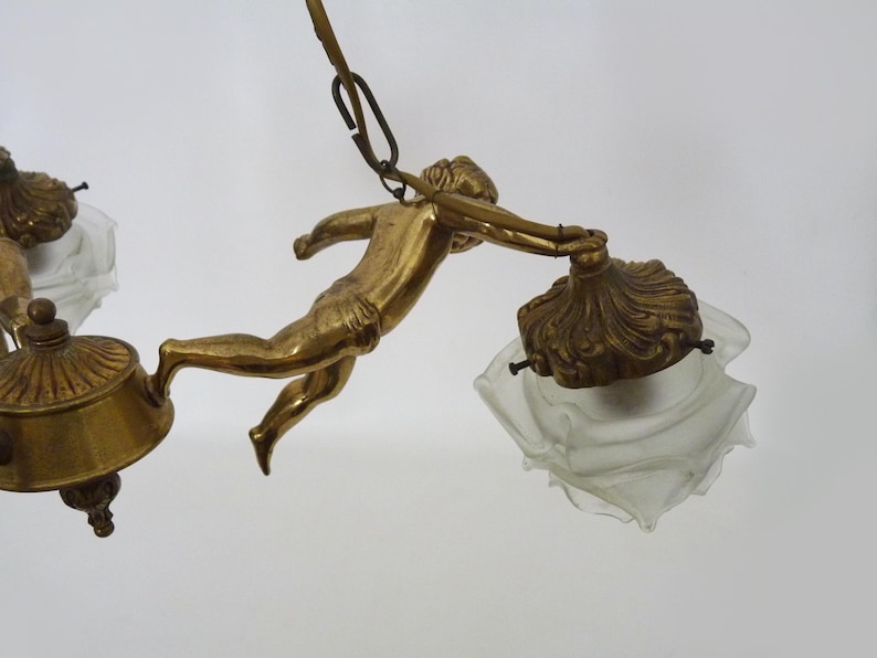 Puede incluir: Candelabro antiguo de lat&oacute;n con una figura de querub&iacute;n que sostiene una l&aacute;mpara. La l&aacute;mpara tiene un dise&ntilde;o decorativo y ornamentado con pantallas de vidrio esmerilado. El dise&ntilde;o general es vintage y elegante.