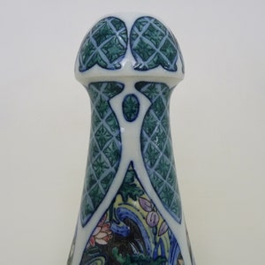 May include: A white ceramic bottle stopper with a green and blue geometric design. The top of the stopper is shaped like a dome with a decorative pattern.