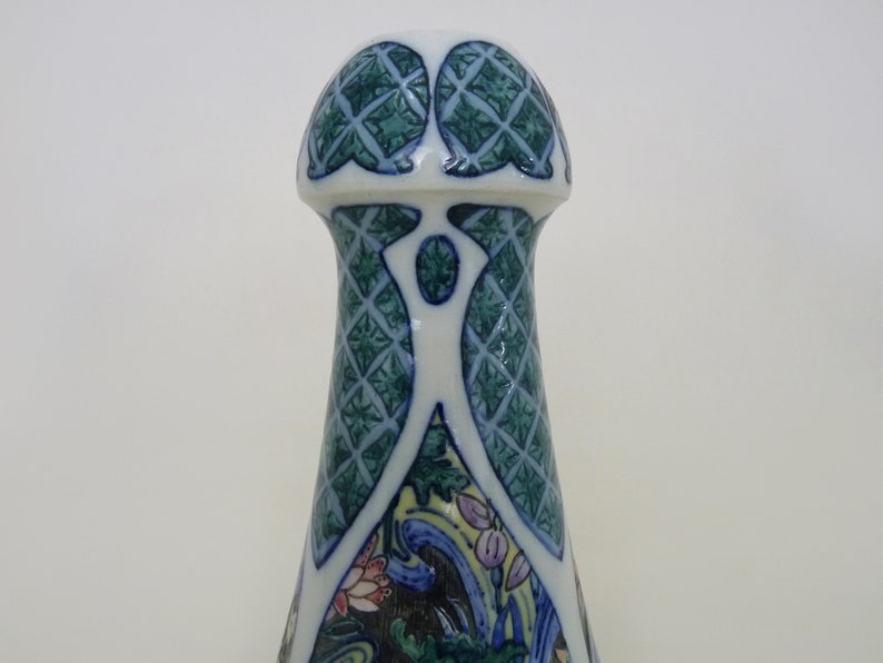 May include: A white ceramic bottle stopper with a green and blue geometric design. The top of the stopper is shaped like a dome with a decorative pattern.