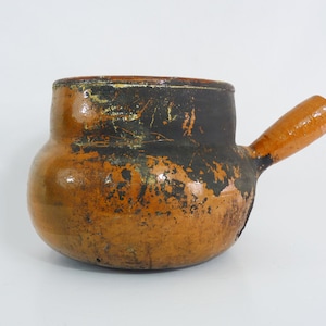 Può includere: Un vaso di ceramica marrone con un manico di legno. Il vaso ha un aspetto usurato e rustico con una smaltatura nera e marrone.