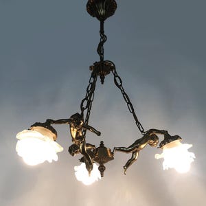 Puede incluir: L&aacute;mpara de ara&ntilde;a antigua de bronce con tres figuras de querubines sosteniendo pantallas de vidrio esmerilado. La l&aacute;mpara est&aacute; suspendida por cadenas y tiene detalles intrincados. Las luces est&aacute;n encendidas, emitiendo un brillo c&aacute;lido.