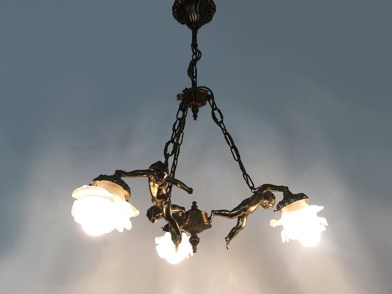 Puede incluir: L&aacute;mpara de ara&ntilde;a antigua de bronce con tres figuras de querubines sosteniendo pantallas de vidrio esmerilado. La l&aacute;mpara est&aacute; suspendida por cadenas y tiene detalles intrincados. Las luces est&aacute;n encendidas, emitiendo un brillo c&aacute;lido.