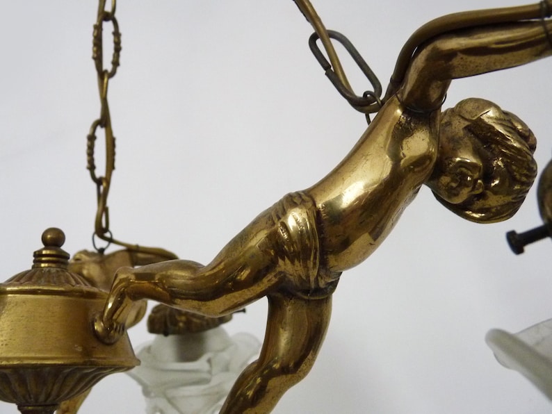 Puede incluir: Primer plano de una l&aacute;mpara de ara&ntilde;a de lat&oacute;n vintage con una figura de querub&iacute;n esculpida. El querub&iacute;n est&aacute; en una pose din&aacute;mica, con detalles intrincados. La l&aacute;mpara de ara&ntilde;a tiene un tono dorado c&aacute;lido y est&aacute; suspendida por cadenas.
