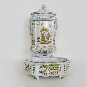 Pode incluir: Fonte de parede em cerâmica branca com tampa, bacia e torneira. A fonte é decorada com cenas florais e pastorais verdes, amarelas e laranja. A bacia tem uma borda ondulada e repousa sobre três pés pequenos.