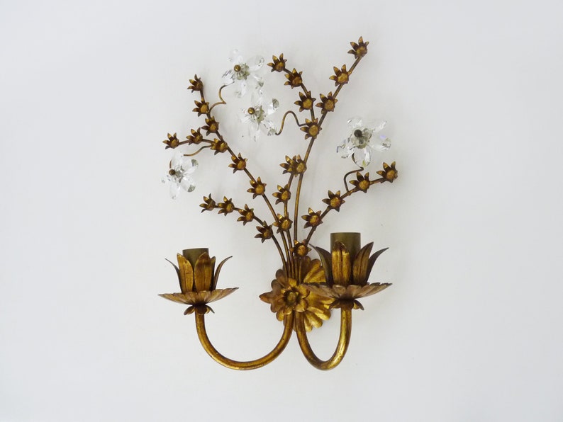 May include: Gold metal wall sconce with a floral design featuring crystal accents. The sconce has two light bulbs and a curved arm.