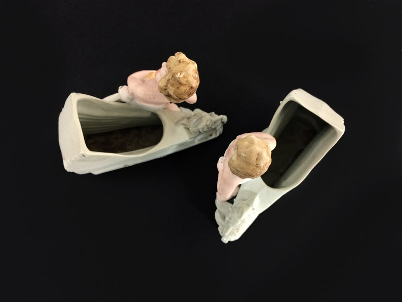 Puede incluir: Dos macetas de cer&aacute;mica blanca con forma de barco con una figura de querub&iacute;n en la proa. Las macetas est&aacute;n vac&iacute;as y tienen un esmalte verde claro.