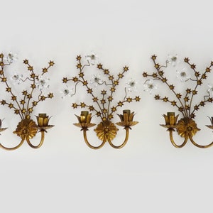 May include: Set of three gold-toned wall sconces with crystal accents. Each sconce features a floral design with delicate branches and leaves. The sconces have two candle-style light bulbs.