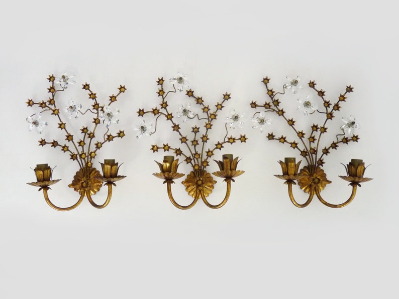 May include: Set of three gold-toned wall sconces with crystal accents. Each sconce features a floral design with delicate branches and leaves. The sconces have two candle-style light bulbs.