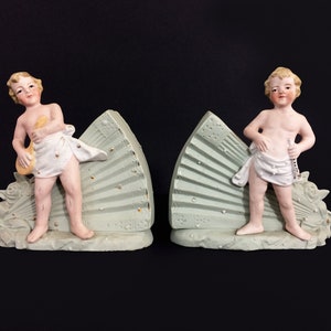 Puede incluir: Un par de sujetalibros de porcelana con figuras de querubines que sostienen instrumentos musicales. Cada querub&iacute;n se encuentra sobre una base en forma de abanico verde y dorado.