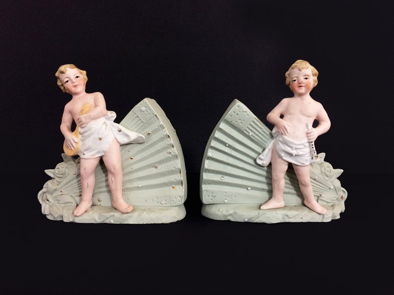 Puede incluir: Un par de sujetalibros de porcelana con figuras de querubines que sostienen instrumentos musicales. Cada querub&iacute;n se encuentra sobre una base en forma de abanico verde y dorado.