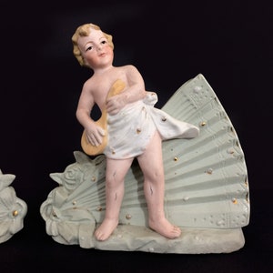 Puede incluir: Un par de figuritas de porcelana de querubines, cada uno sosteniendo un instrumento musical, de pie sobre una base en forma de abanico verde y dorada.