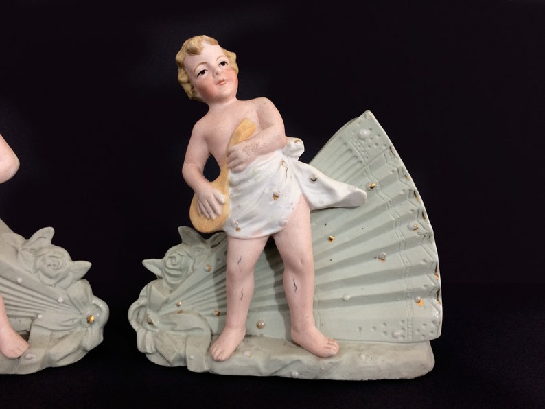 Puede incluir: Un par de figuritas de porcelana de querubines, cada uno sosteniendo un instrumento musical, de pie sobre una base en forma de abanico verde y dorada.