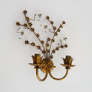 May include: Gold metal wall sconce with a floral design featuring crystal accents. The sconce has two light bulbs and a gold finish.