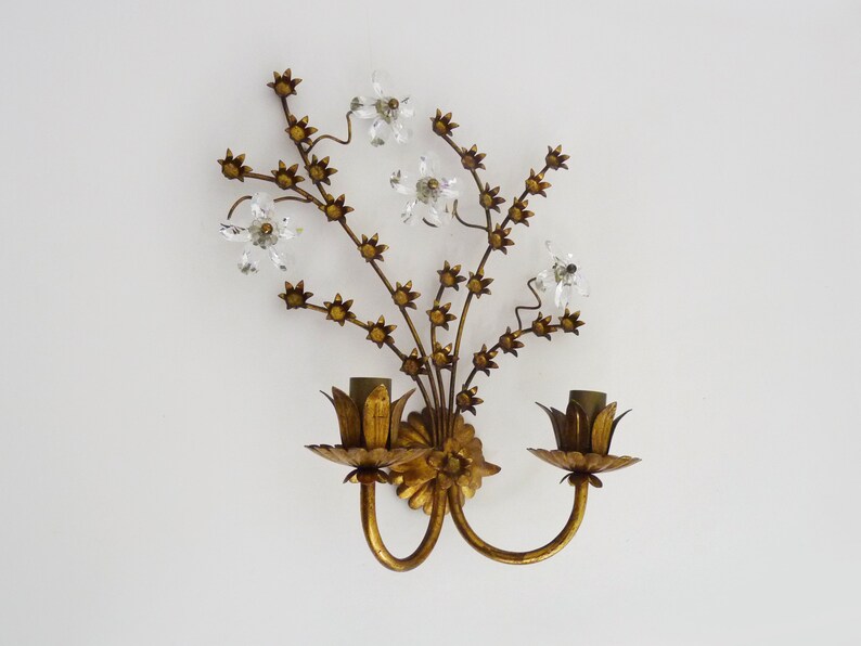 May include: Gold metal wall sconce with a floral design featuring crystal accents. The sconce has two light bulbs and a gold finish.