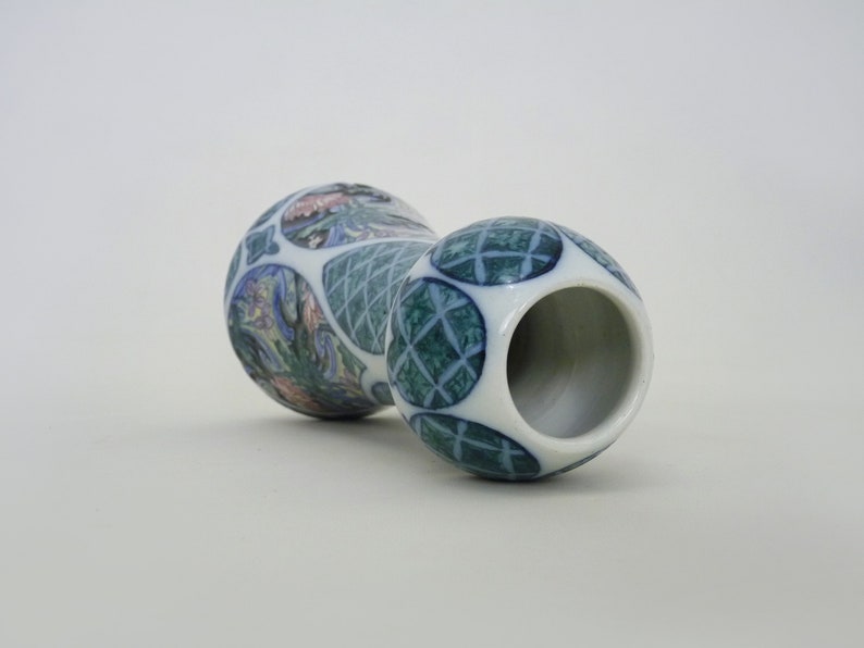 May include: A white ceramic vase with a blue and green geometric pattern and a floral design. The vase has a wide mouth and a narrow base.