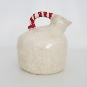 May include: A cream-coloured ceramic jug with a red and white striped handle. The jug has a rounded body and a spout. The handle is arched and the jug appears to be handmade.