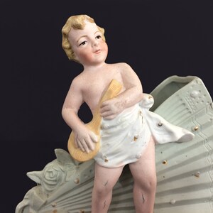 Puede incluir: Una figurita de porcelana de un querub&iacute;n tocando un instrumento musical, de pie sobre una base decorativa en forma de abanico. El querub&iacute;n lleva una tela blanca y tiene el pelo rubio. La base est&aacute; pintada en tonos verdes y dorados.