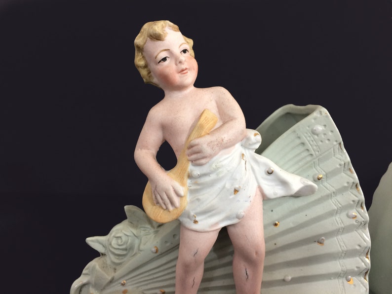 Puede incluir: Una figurita de porcelana de un querub&iacute;n tocando un instrumento musical, de pie sobre una base decorativa en forma de abanico. El querub&iacute;n lleva una tela blanca y tiene el pelo rubio. La base est&aacute; pintada en tonos verdes y dorados.