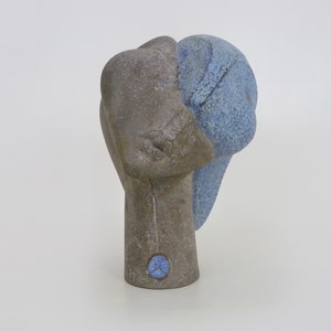 Puede incluir: Una escultura de cerámica de una cabeza con un esmalte azul. La cabeza está inclinada hacia un lado y la cara está parcialmente oscurecida por el esmalte. La escultura está sobre un fondo blanco.