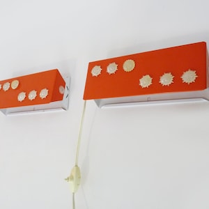 Peut inclure: Deux appliques murales orange avec des accents décoratifs blancs et dorés. Chaque applique a une forme rectangulaire avec une base argentée et un cordon avec une prise. Les appliques sont montées sur un mur blanc.