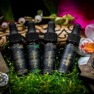 May include: Four small black bottles with gold lettering on the labels. The bottles are arranged in a row on a bed of green moss. The labels read "Valdhalla", "Yggdrasil", "Primal", and "The Monk".