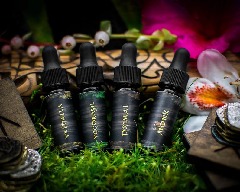 May include: Four small black bottles with gold lettering on the labels. The bottles are arranged in a row on a bed of green moss. The labels read "Valdhalla", "Yggdrasil", "Primal", and "The Monk".