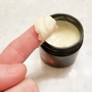 Puede incluir: Primer plano de un dedo que se introduce en un peque&ntilde;o tarro negro de crema blanca. La crema es espesa y tiene una textura suave.