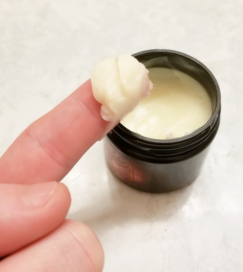 Puede incluir: Primer plano de un dedo que se introduce en un peque&ntilde;o tarro negro de crema blanca. La crema es espesa y tiene una textura suave.