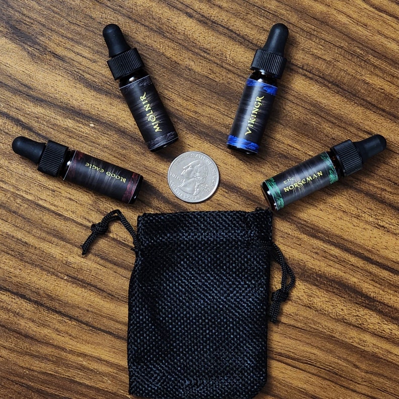 May include: Four small glass bottles with black labels and gold lettering. The bottles contain essential oils or perfumes. The labels read "Blood Eagle", "The Norsemen", "Mjolnir", and "Vikingr". A black drawstring bag is in the center of the image.