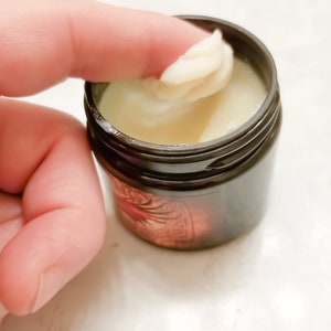 Puede incluir: Un peque&ntilde;o tarro negro con una sustancia blanca y cremosa en su interior. Un dedo se introduce en el tarro y saca un poco de la crema.