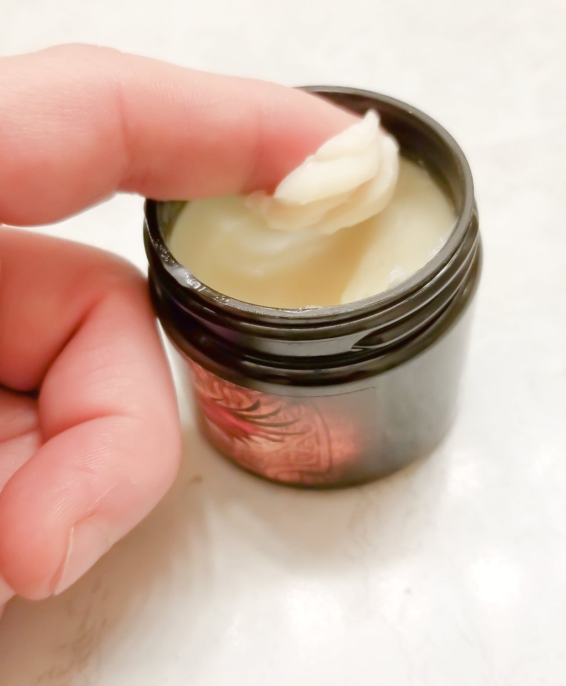 Puede incluir: Un peque&ntilde;o tarro negro con una sustancia blanca y cremosa en su interior. Un dedo se introduce en el tarro y saca un poco de la crema.